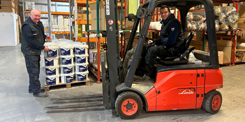Two warehouse operatives, one stood next to tubs of adhesives and the second sat on a forklift truck