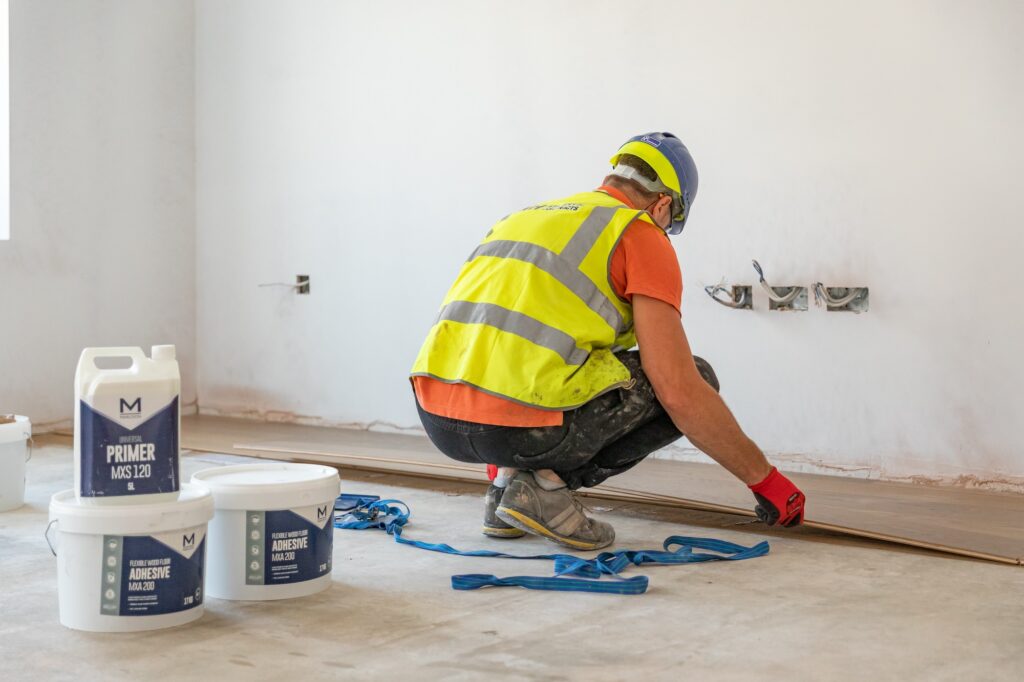 This image shows a site-worker in a high-vis kneeling down, facing away from the camera to lay wood flooring and to the left of him, are two tubs of Marldon MXA200 wood floor adhesive and 1 bottle of MXS120 universal primer.