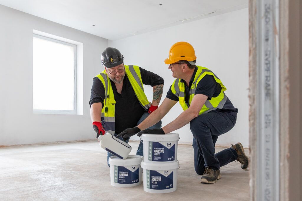 This image shows two site-workers kneeling in the middle of a recentl plastered room, they are both wearing high-vis vests, one is looking down at 3 tubs of Marldon MXA200 adhesive and other is looking towards him.